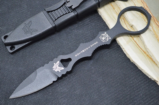 An image featuring the Benchmade 177BK SOCP dagger, which has a black combat fixed spear point blade and molded plastic handle. The dagger is displayed on a black and wood surface. Next to it is the accompanying sheath.